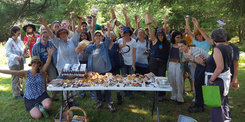 Participants at a Catskill Fungi mushroom walk gathered around a table covered with foraged mushrooms, and holding up mushrooms jubilantly.