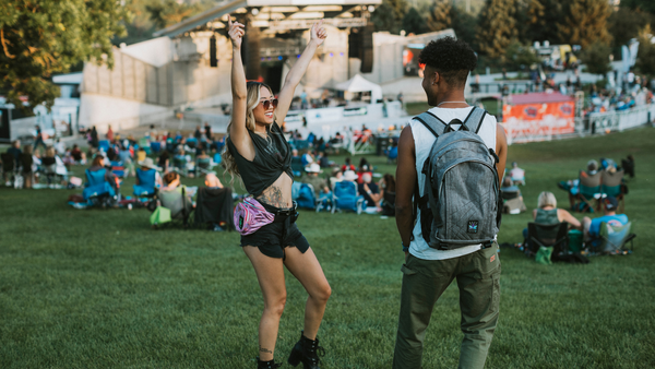 Girl with fanny pack dancing at music festival while she is watched by her friend wearing a backpack.