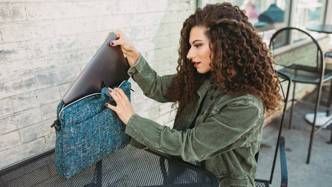 Girl with curly hair and olive green jacket sitting outside putting Macbook computer into Glass Side Hustler