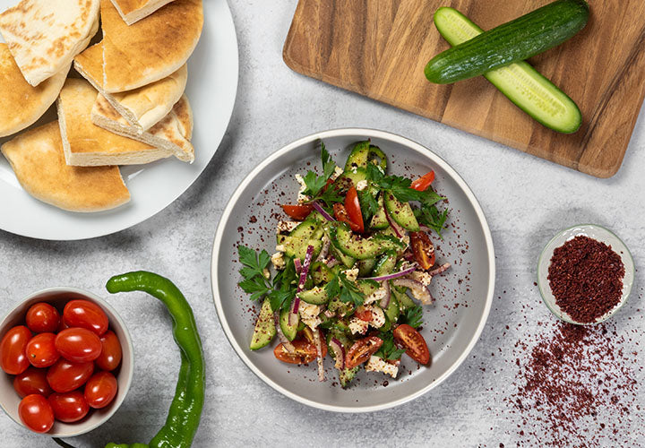 Turkish salad of cucumber, onion, wax pepper, parsley, tomato, and feta cheese on a plate with sumac spice and pita bread.