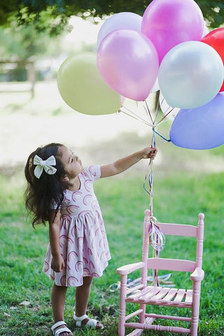 toddler in rainbow dress with balloons tied to little pink chair
