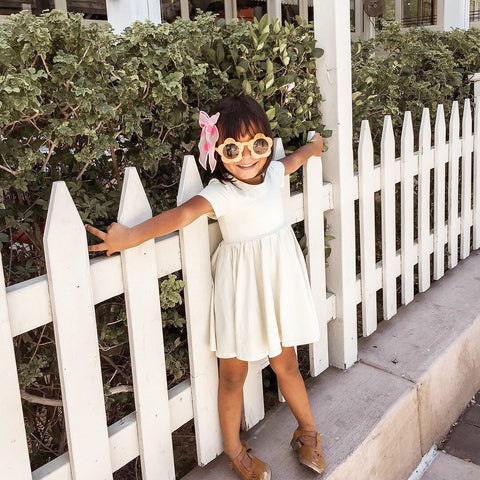 cute kid in yellow dress leaning on white picket fence wearing flower shaped sunglasses