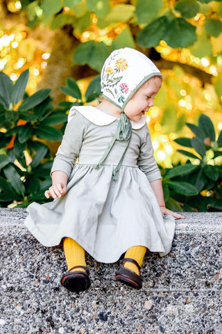 toddler in green twirl dress, yellow tights and embroidered bonnet sitting on bench