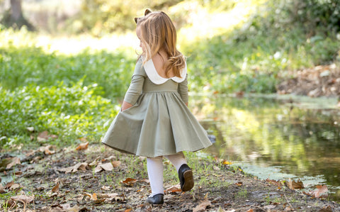 toddler walking near a creek with hands in pockets of her green fall dress