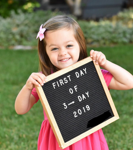 three year old girl holding first day of school sign in pink dress and pink bow