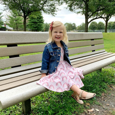 toddler girl sitting on park bench with jean jacket and rainbow dress