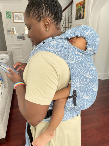 A black mama wears her black baby on her back in 100% linen Lotus Blue hybrid half buckle baby carrier. She is working on her phone while baby sleeps with the hood up cradling his head.