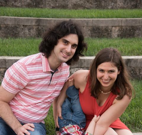 Alexandra, Founder of Mama & Roo's Supplement Co, with husband. Young white couple smiling, sitting on a grassy step at a park, holding a child upside down between them. Child's face is not shown for privacy