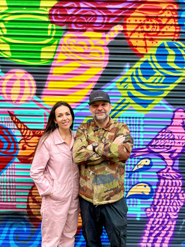 artist and wife standing infront of a mural in devon, a street a mural made with spray paint and brightly coloured