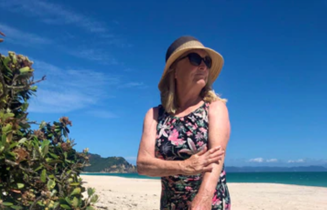 middle aged woman wearing a hat and sunglasses rubbing her arm at the beach with tree next to her