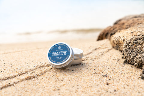 two tins of Seasick Sunscreen on a sandy beach next to a log
