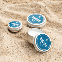 Three tins of Seasick Sunscreen in various sizes sitting on the sand one with with lid off