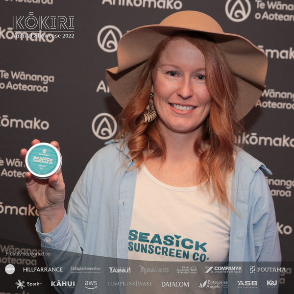 woman wearing hat holding tin of Seasick Sunscreen while smiling standing in front of a black branded background