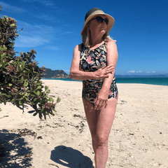 woman in her sixties wearing a hat and swimsuit rubs sunscreen onto her arm at the beach