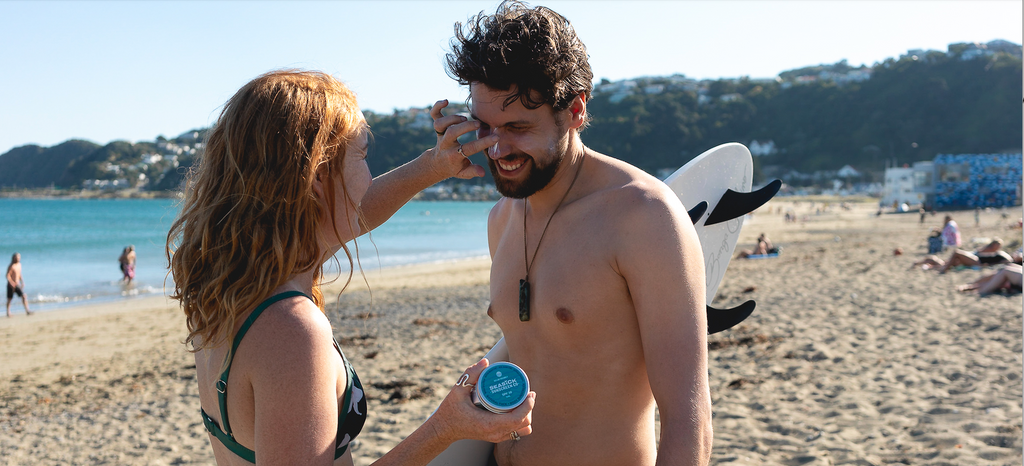 woman with red hair applies sunscreen to man's nose carrying surfboard at the beach