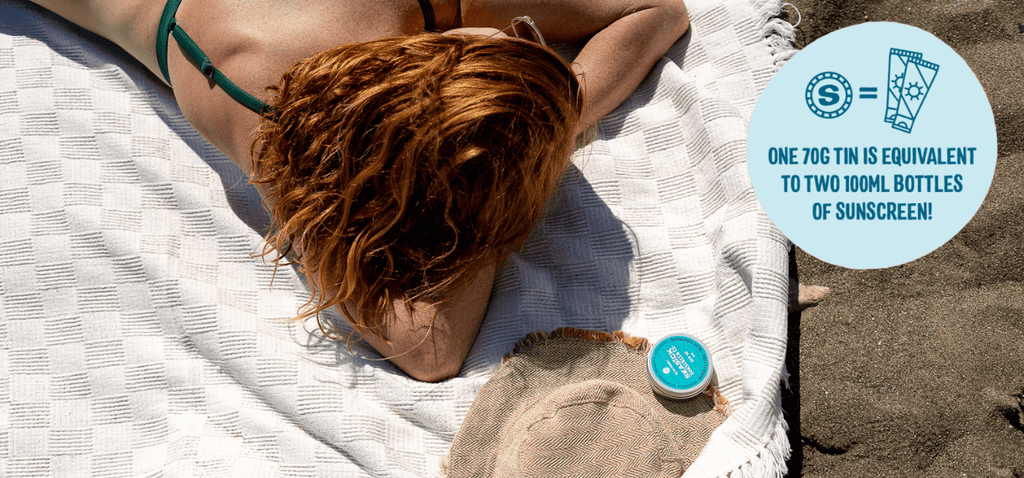 woman with red hair sunbathing on a white beach towel with a tin of sunscreen and a hat