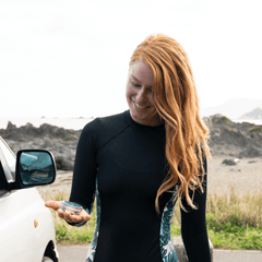 woman with red hair in a surf suit looking at a tin of sunscreen in her palm