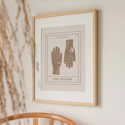 A neutral coloured print, framed hanging on the wall, featuring two hand illustrations side by side on a tarot card. The initials of the couple are shown on each wrist, with a special personalised date at the top. Below the hands it says 'The Lovers' in capital text.