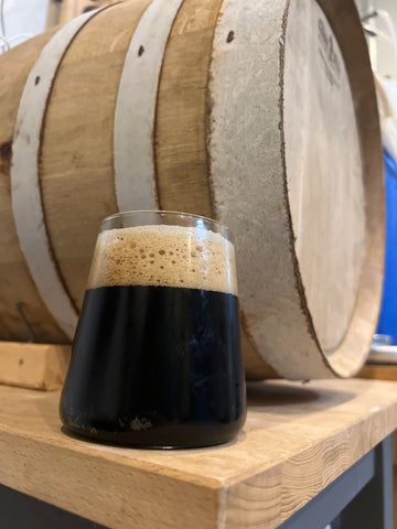 Glass of Schwarzbier placed next to a barrel at Oil Creek Brewing