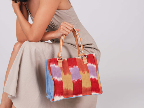 A model holds the Mini Irma Tote in Raspberry Paleta pattern. It has a watercolor red, orange, yellow, purple, and white stripes. The sides are sky blue. It has leather handles with white embroidery.