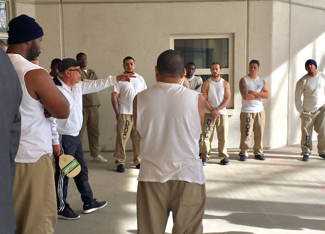 Roger BelAir stands with a group of male inmates. He holds a pickleball paddle while he instructs the group.