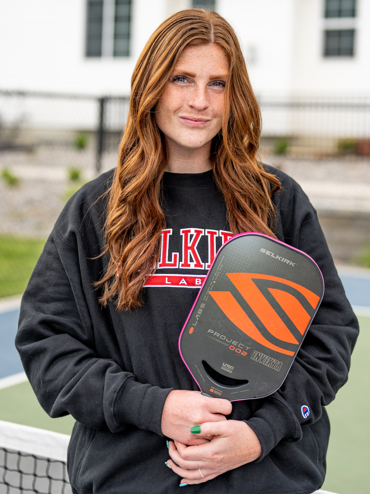 Selkirk Emerging Pro Brooke Bromley stands on a pickleball court near the net. She holds her Project 002 paddle and smiles at the camera.