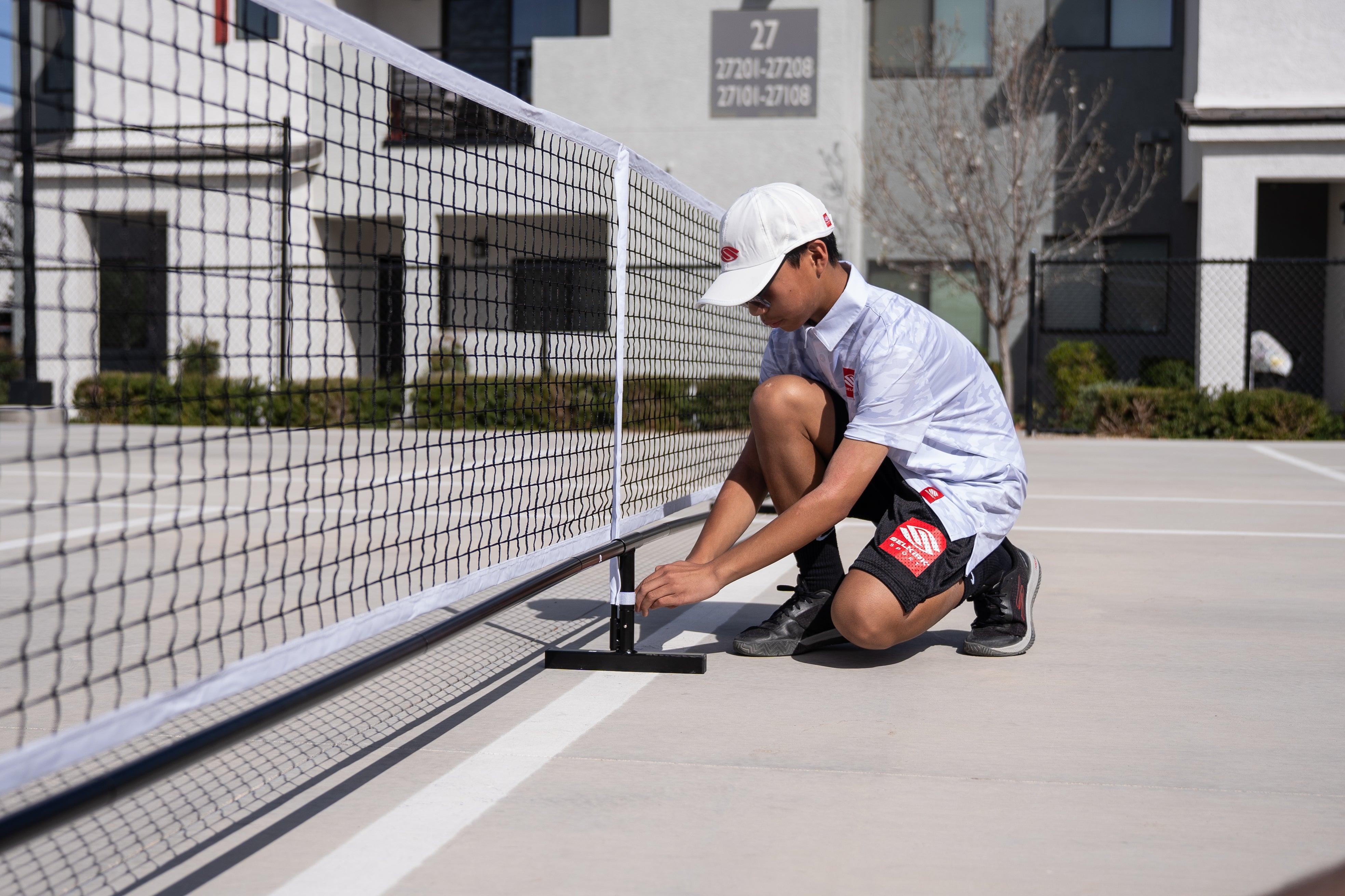 A man sets up a temporary pickleball net on a pickleball court. He squats down to attach the center post to the net.