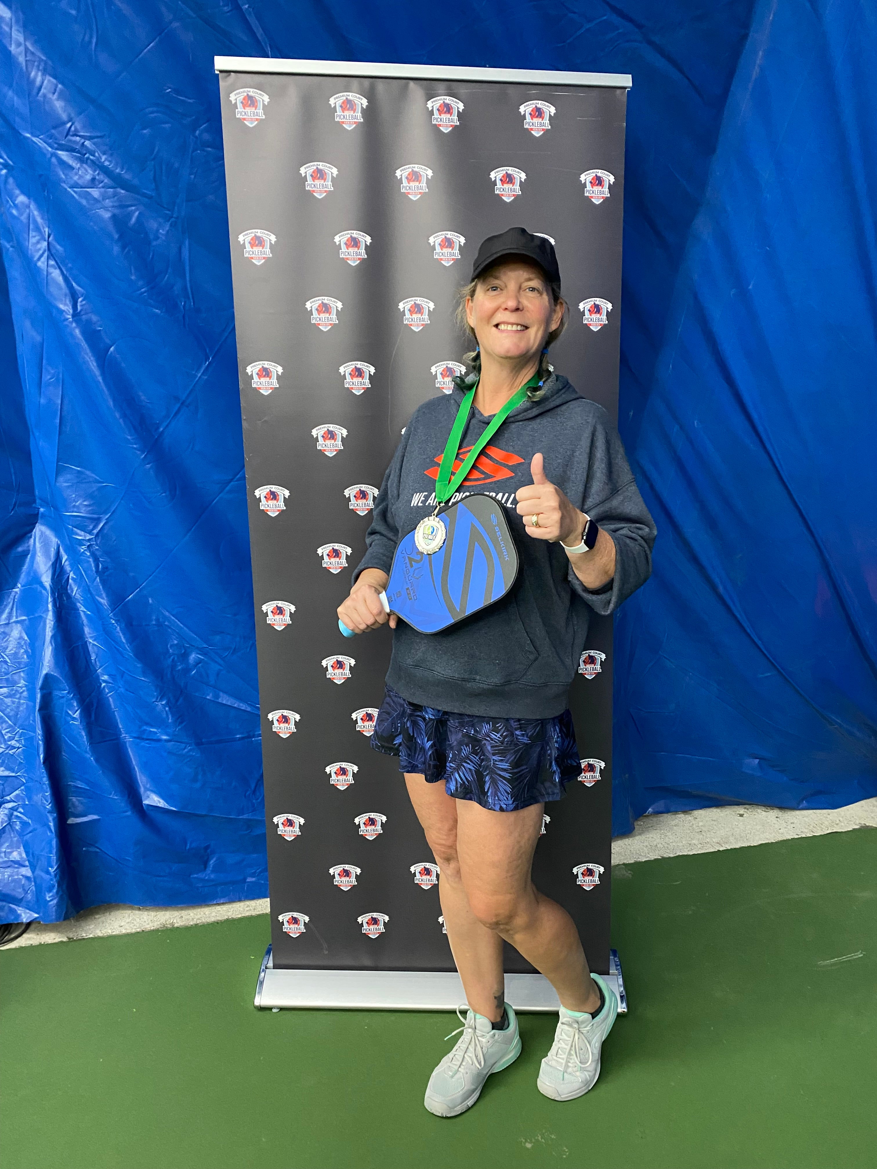 Kelli McRobert gives a smile and thumbs up to the camera. She is holding her pickleball paddle and wearing a gold medal.