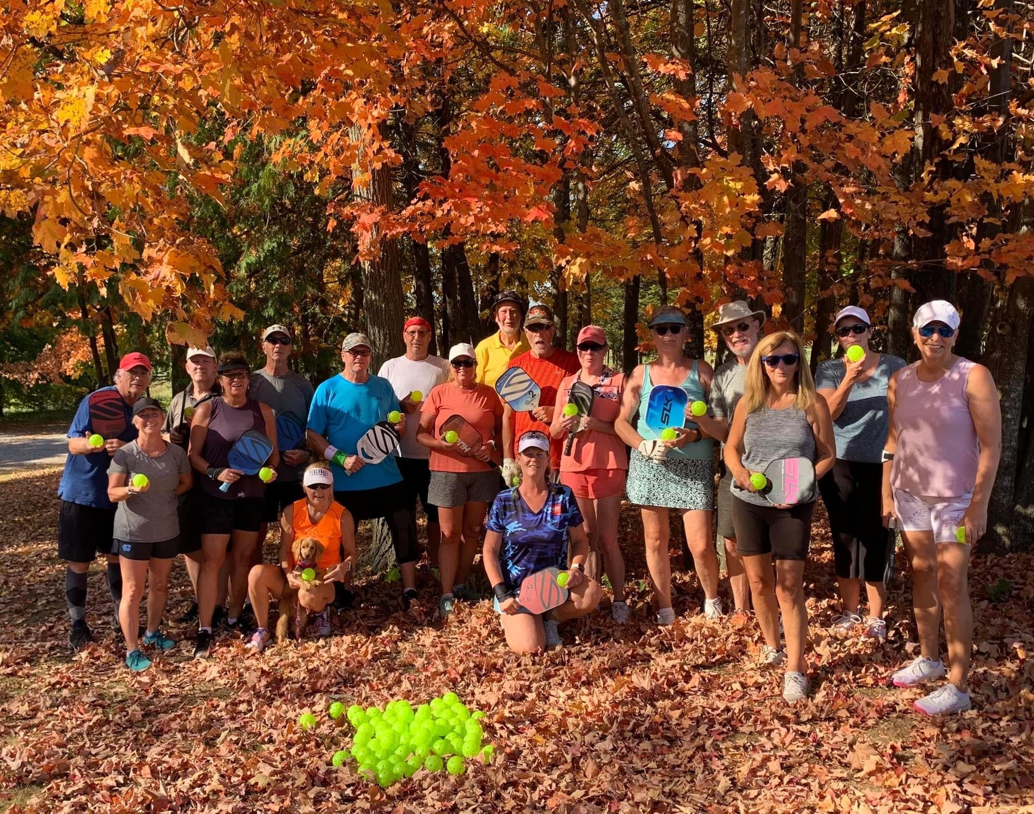A group of pickleball players stand in fall foliage with their pickleball paddles.