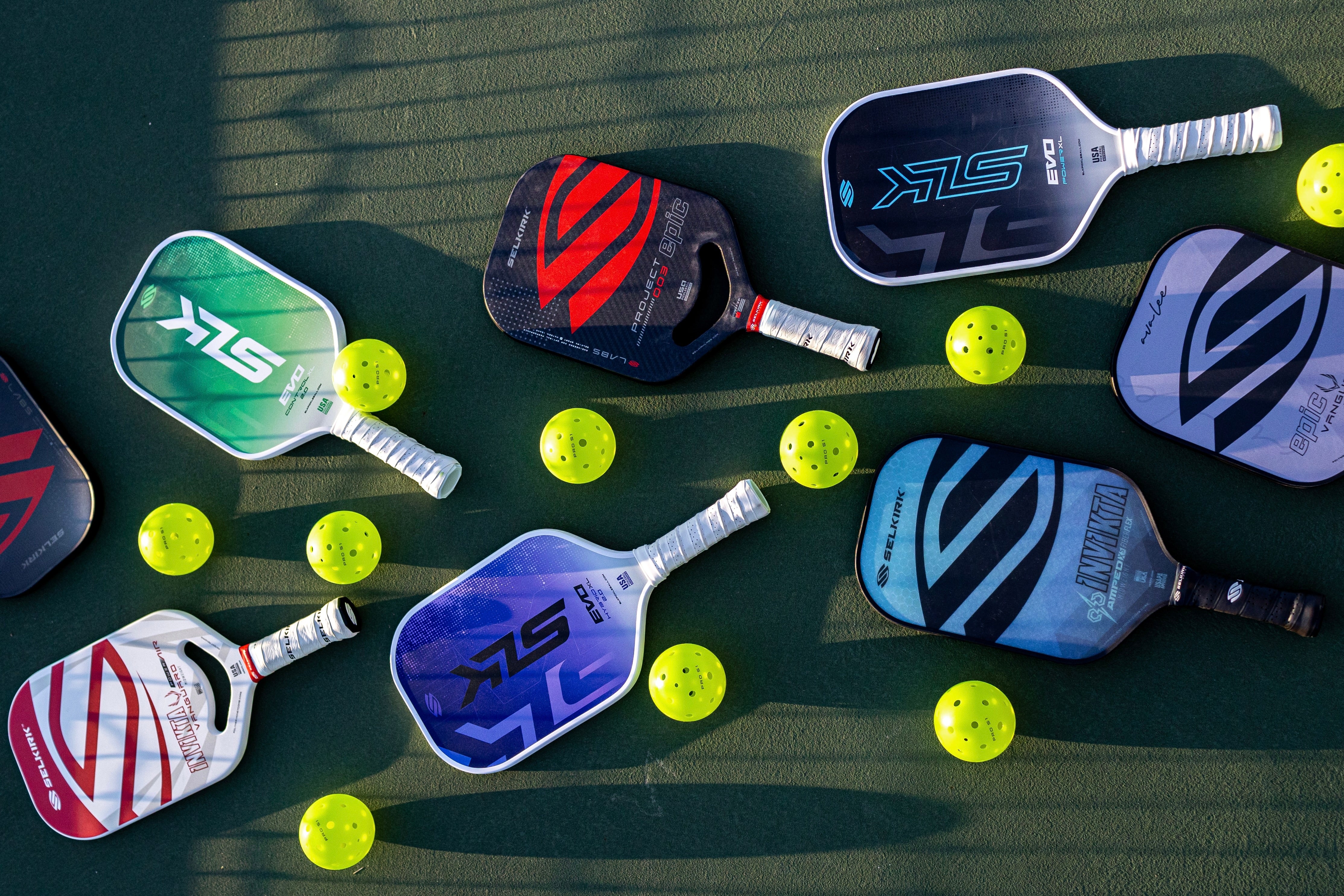 An array of Selkirk Sport pickleball paddles lay face up on a pickleball court. Scattered around them are several pickleballs.