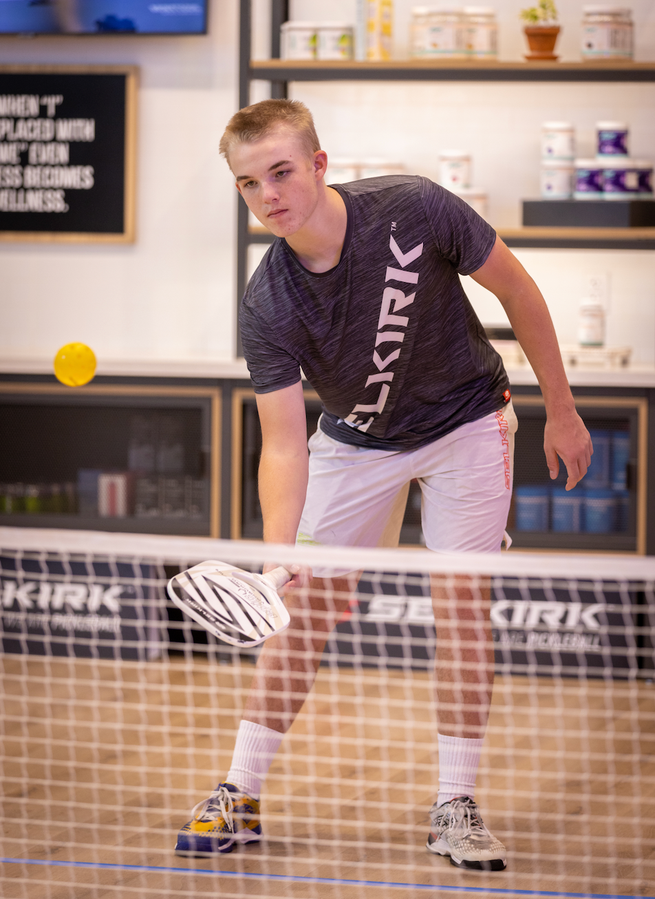 A teenage boy plays pickleball indoors. He stands on a wooden floor ready to hit a ball at the net.