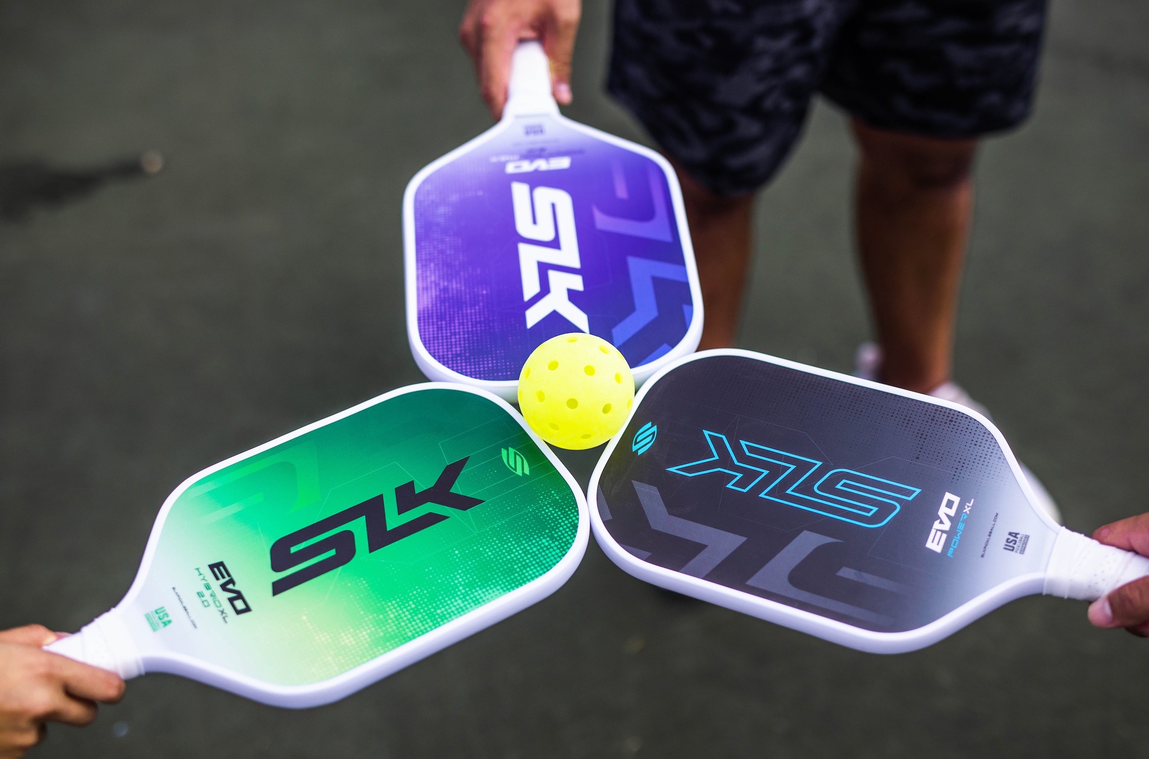 Three players hold their SLK Evo 2.0 paddles in a triangle shape. The paddles are touching and balanced in between all three is a pickleball.