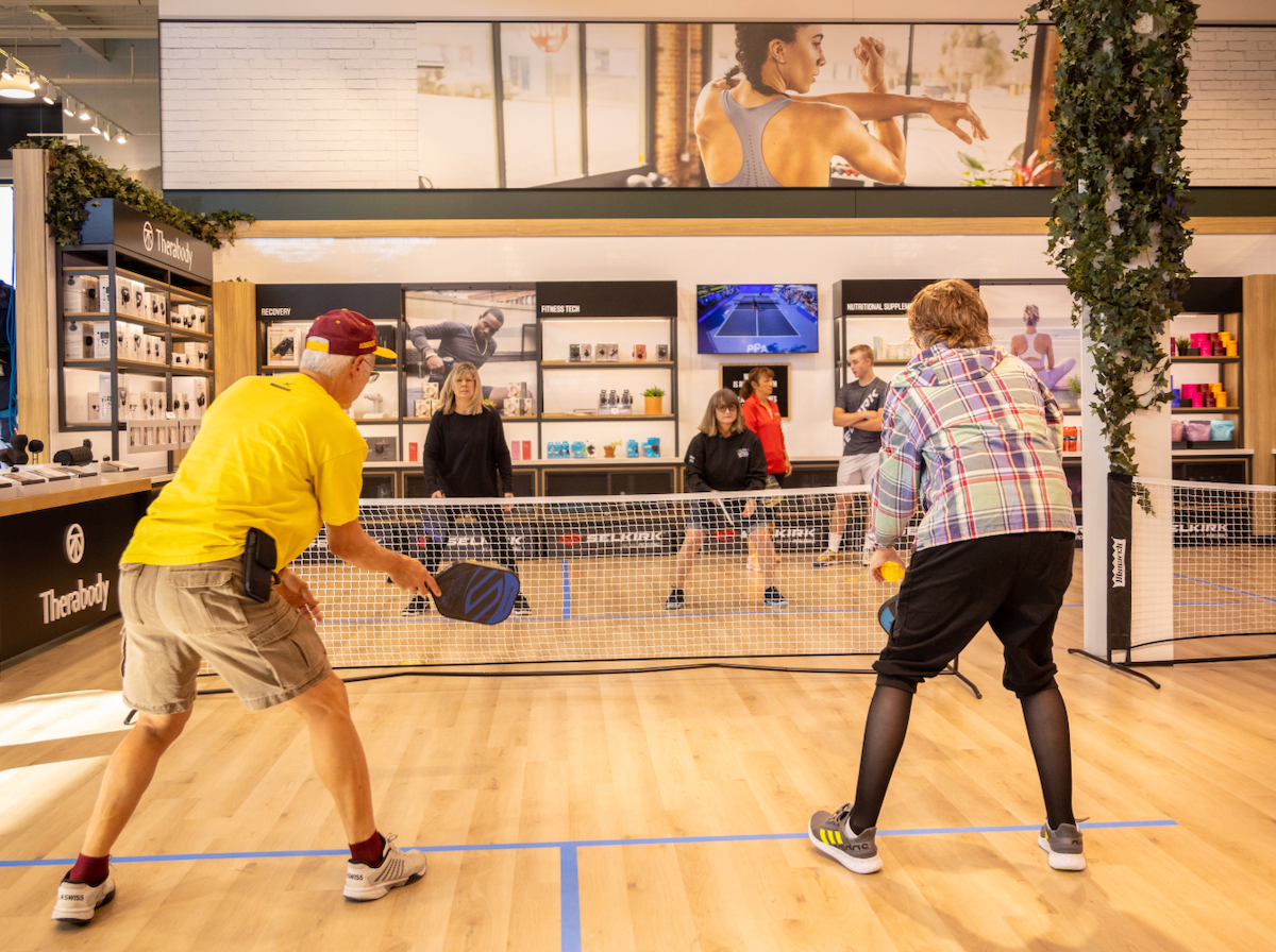 Four people play pickleball on a court inside a DICK's Sporting Good store. A man and woman stand with their backs to the camera, ready to receive a ball from the other team.