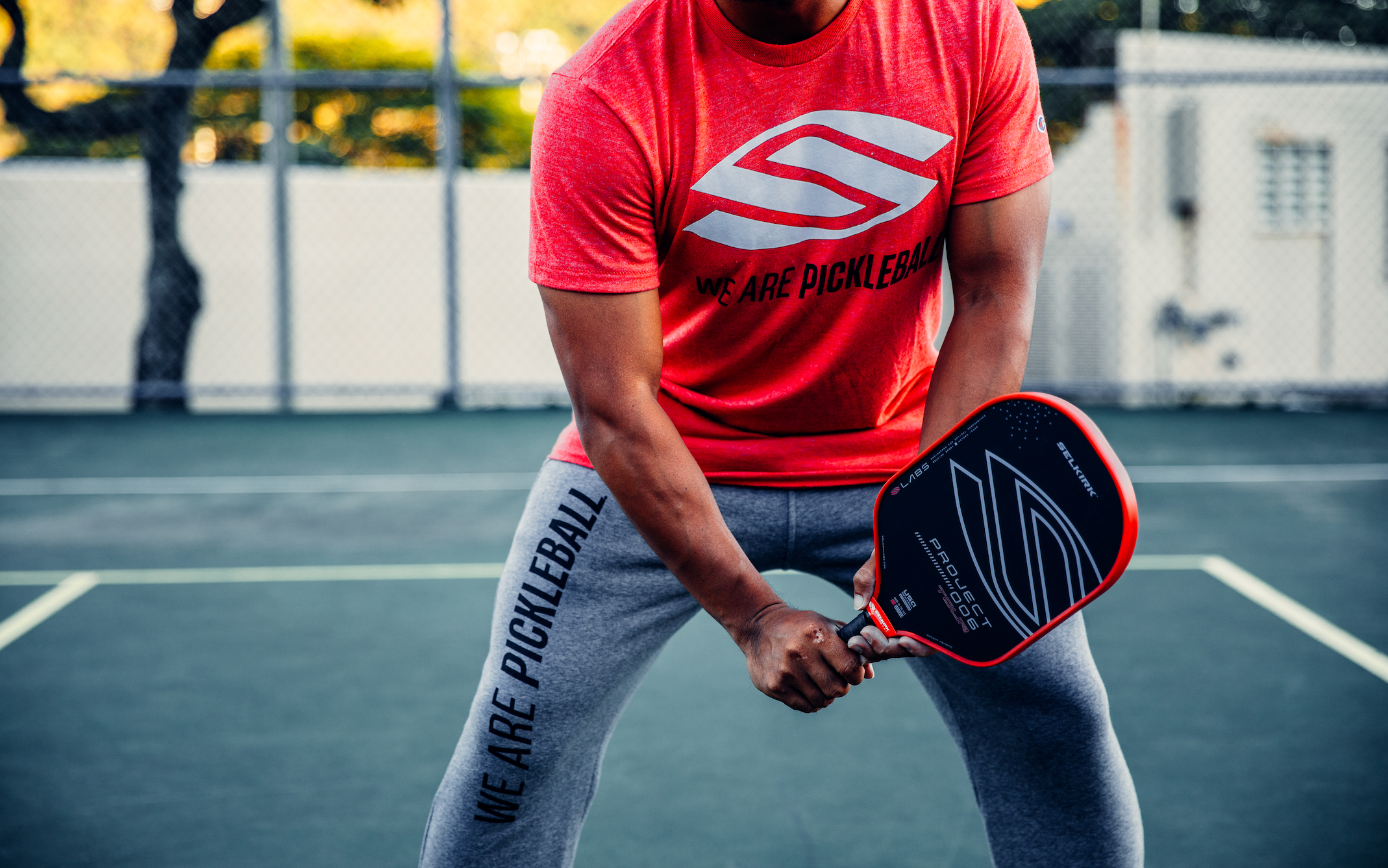 A man stands at the kitchen line of a pickleball court. He leans to his left with his Selkirk Project 006 paddle in front of him.