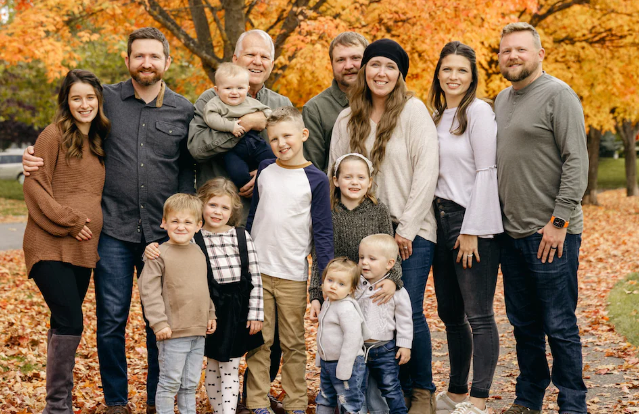 The entire Barnes family stands together in a park. They are smiling at the camera and behind them is fall foliage.