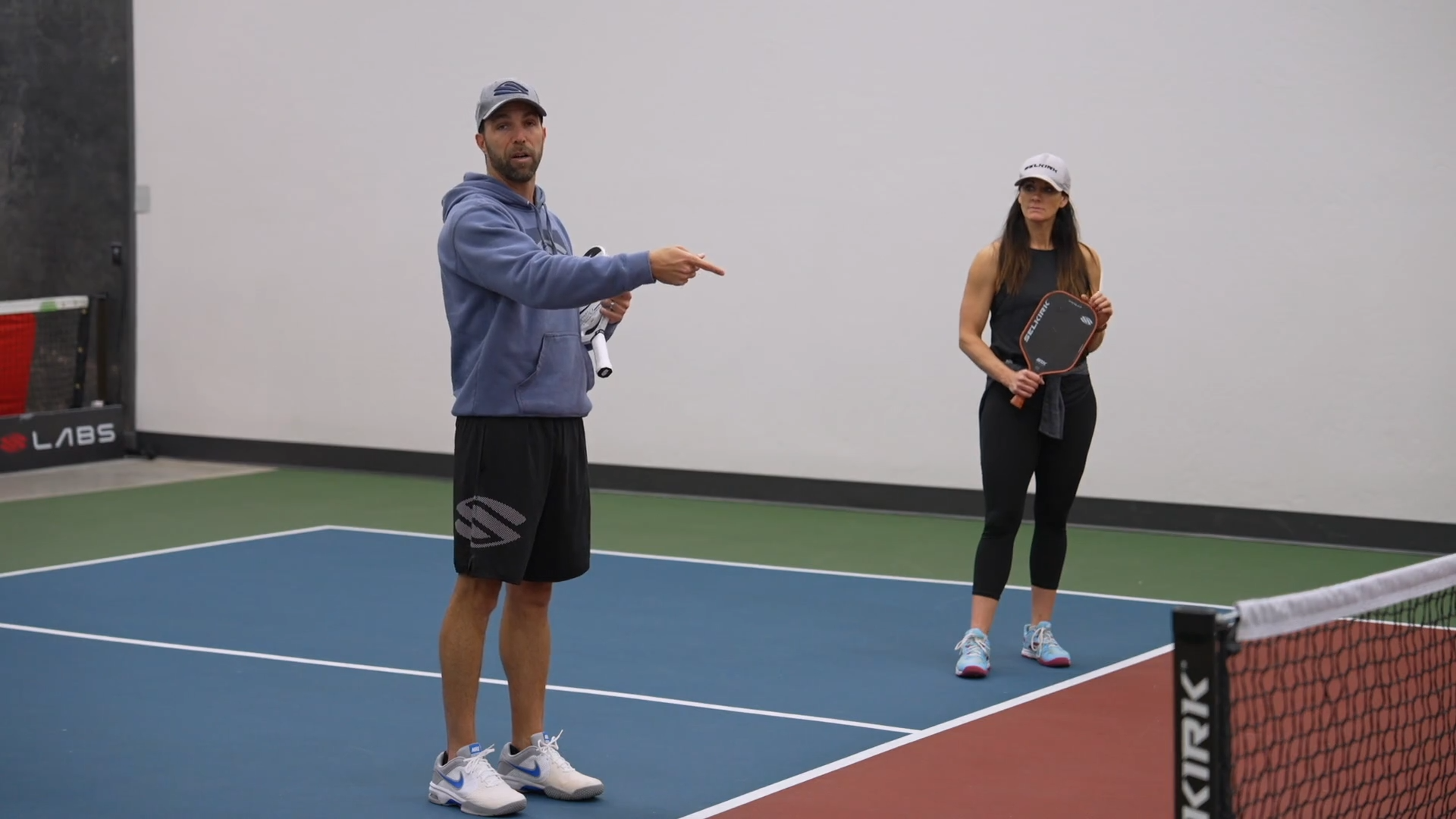 Daniel J Howard stands on an indoor pickleball court with his arm outstretched. He is teaching a woman, who stands on the same side of the court, a new pickleball skill.
