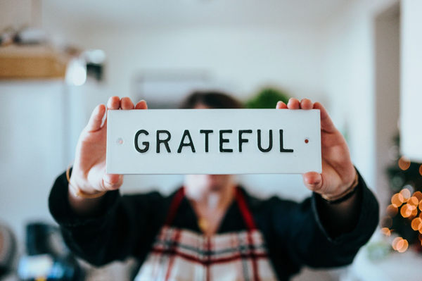 A person holding up a sign that reads the word "grateful." The sign is blocking the persons face. The person is wearing an apron and is in the kitchen.