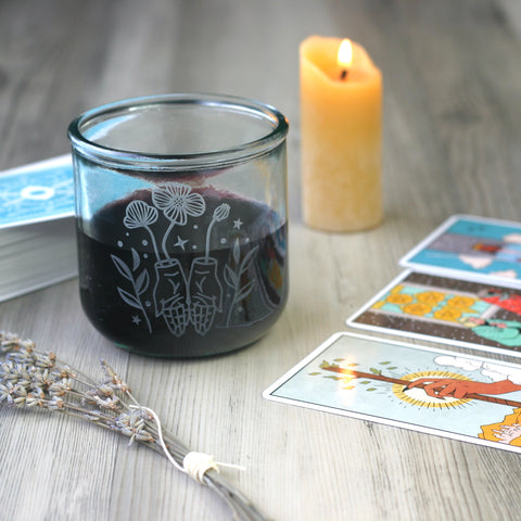 Glass tumbler with an engraved design of a pair of hands held open, palms up, with poppy flowers growing out of the wrists. There are simple leaf stalks and stars surrounding the hands and flowers. The glass has red wine in it, and sits near Tarot cards, a candle, and a sprig of lavender.