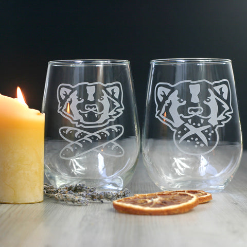 A pair of wine glasses sitting on a table next to a lit beeswax candle. There are graphics engraved on each glass of American badger faces. One is smiling above a pair of crossed baguettes, and the other has a round pretzel roll in its mouth.