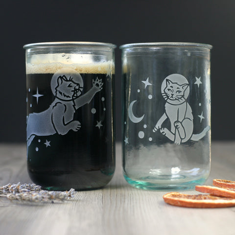 a pair of glass tumblers with astronaut cats engraved on them. The cats both have bubbles around their heads, and are surrounded by stars. One is stretched out in a leap, reaching for a star. The other cat is dangling uncomfortably and its eyes are slightly crossed.