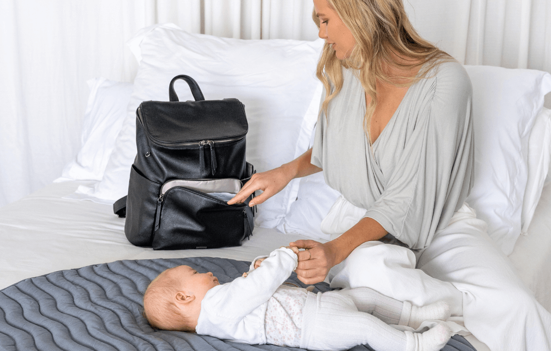 mum and newborn baby sit on bed. Mum is taking wipes out of a stylish black baby bag made from vegan leather.