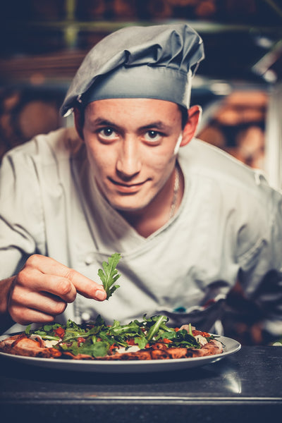 Chef looking towards the camera while topping a pizza with herbs.