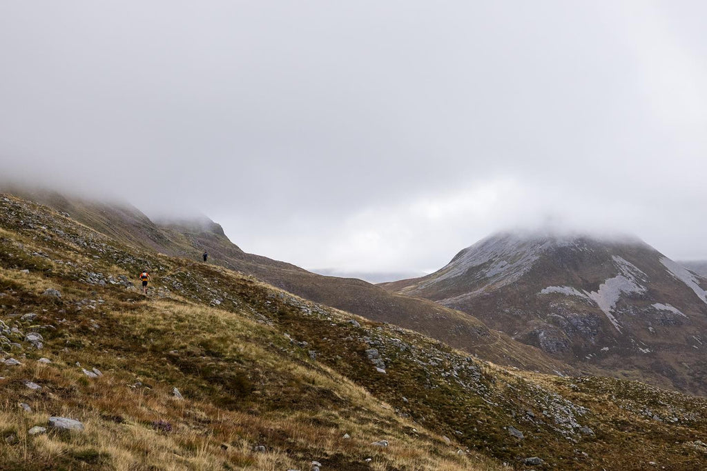 Runners in the distance struggle over rough terrain on a bleak, cloudy Scottish landscape.