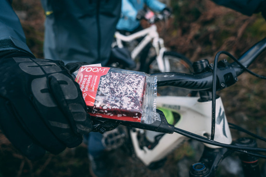 A gloved hand holds a VOOM Beetroot energy bar above the handle bars of a mountain bike during a mid-ride snack stop