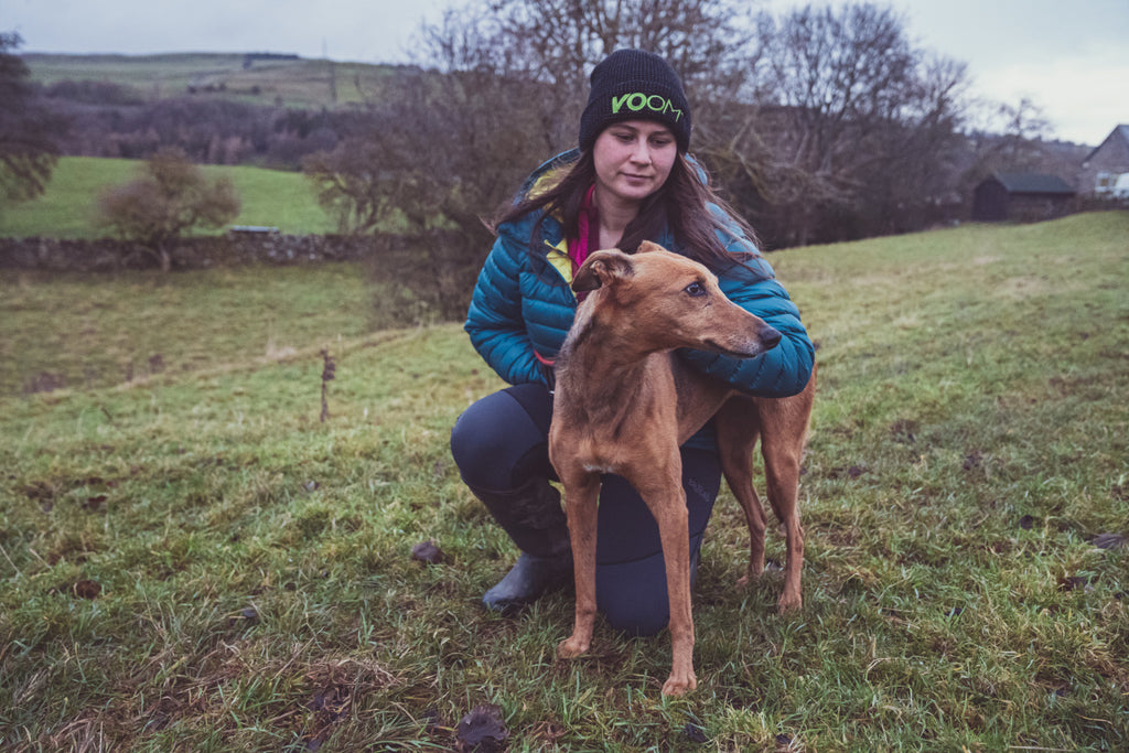 A lady wearing a winter jacket and VOOM beanie hat kneeling down next to her dog