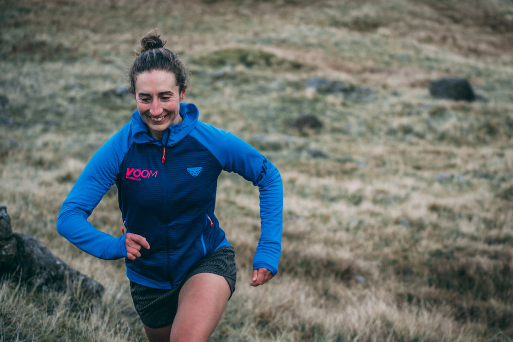 A female runner with a big smile on her face strides out up a grassy hill