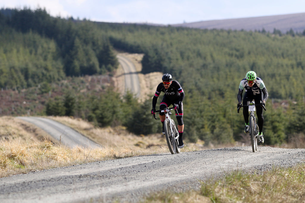 Two cyclists sprint up a hill during a gravel ride with a large expanse of forest behind them