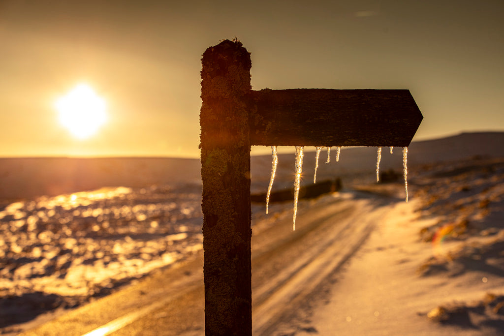 A golden sun illuminates a snowy fell side scene with icicles hanging from a footpath sign post
