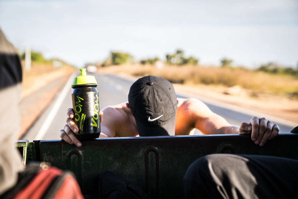 A male runner leans on the back of a vehicle, water bottle in hand under a hot sun