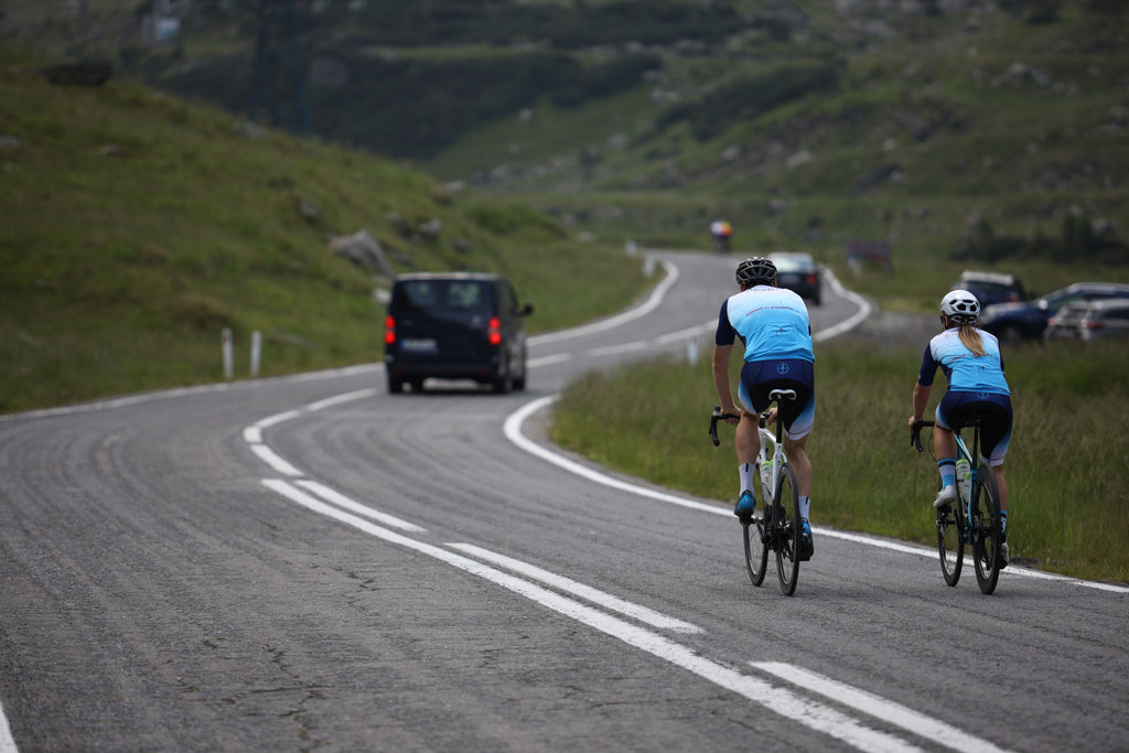 A male and female cyclist ride up a hill following a van